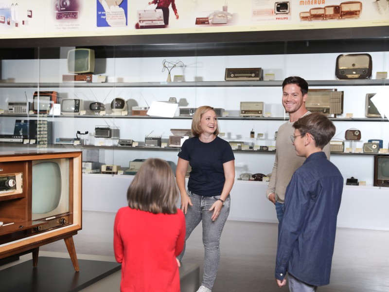 A cultural mediator with a family in the exhibition medien.welten, in the background historical television sets: 